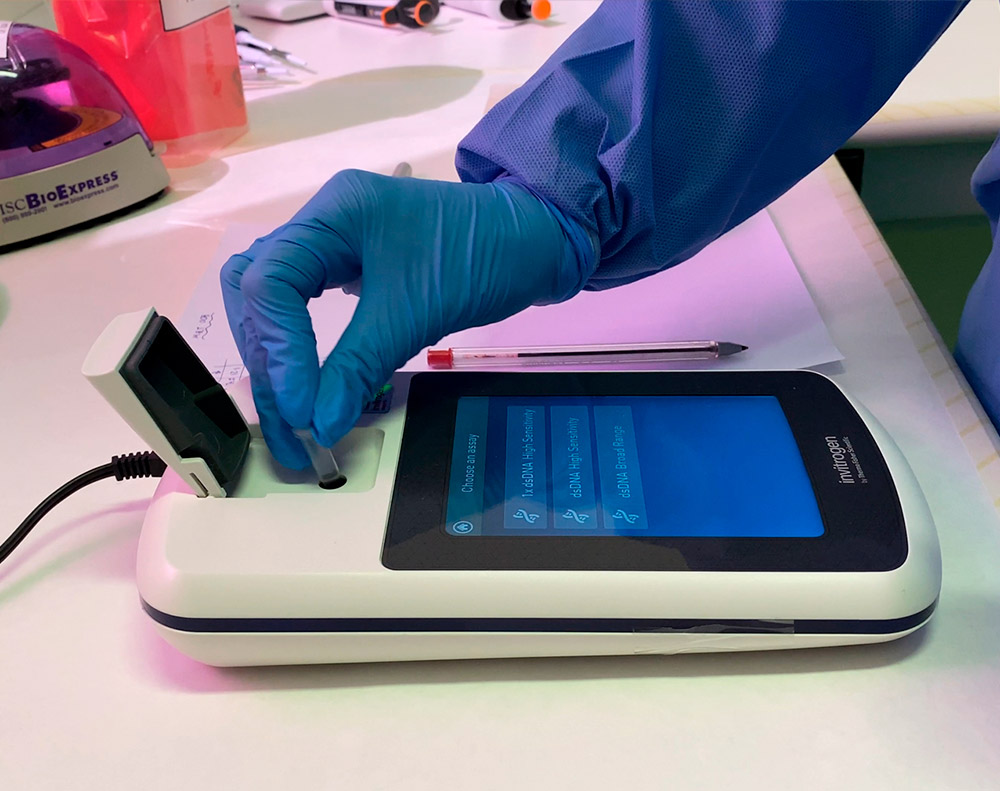 Fotografia retratando um dos procedimentos na rotina de um laboratório de pesquisa que faz análises com material biológico. Sobre uma bancada de dor branca, temos um aparelho denominado fluorômetro, que mede a fluorescência emitida em uma amostra depositada dentro de um tubo específico. O fluorômetro em questão consiste em um aparelho retangular com as bordas arredondadas, em cor branca e com uma tarja azul escura. Emoldurada por um retângulo preto, temos uma tela que exibe um menu em cor azul com algumas opções que não estão legíveis na foto. Ao lado direito da bancada, temos a porção do ventre de uma pessoa com avental descartável na cor azul, luvas descartáveis na mesma cor, da qual estão também na fotografia duas mãos. Na mão direita da pessoa, temos um tubo, que está sendo depositado no fluorômetro, em um poço designado para esta função. Próxima a este poço, temos uma tampa com dobradiça, que é parte do aparelho. Ao lado do fluorômetro, temos uma folha de papel e uma caneta, com uma ponta plástica vermelha em uma das extremidades, e a ponta esferográfica prateada na outra extremidade. Sobre a bancada, mais ao fundo, também estão alguns cabos de energia, uma microcentrífuga, algumas micropipetas e outros utensílios de laboratório.