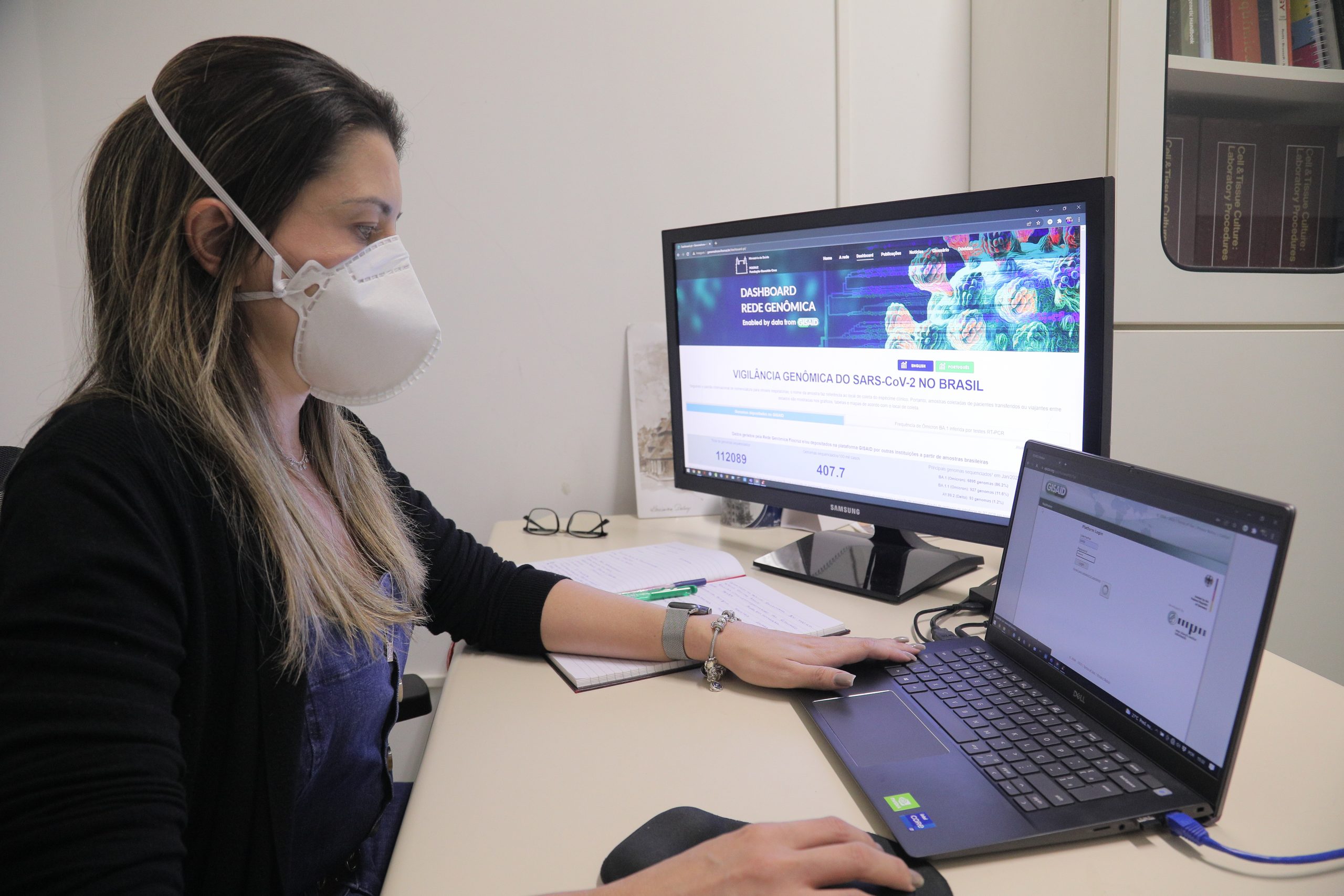 Fotografia ilustrando pesquisadora em processo de análise de dados utilizando computadores. À esquerda da imagem, temos uma mulher de pele bege clara, utilizando máscara facial do tipo PFF2, com roupas comuns — ou seja, sem equipamentos de proteção, uma vez que não está lidando diretamente com a bancada. Ela está vestida com uma camisa azul, e uma blusa preta por cima, com as mangas puxadas até a altura da base do antebraço. A mulher possui ainda cabelos castanho escuros com as pontas descoloridas, usa um esmalte em cor cinza-amarronzada e, no braço esquerdo, um relógio de pulso com pulseira em cor prata. A pesquisadora está sentada a uma mesa, e apenas seu busto está enquadrado na imagem. Sobre a mesa, temos um computador do tipo notebook e um monitor de computador grande, no qual são exibidos dois gráficos, um de barras e outro de área. No notebook está disposta a página do GISAID, o banco de dados internacional cooperativo para depósito e análise de sequências de genoma de Influenza e do SARS-CoV-2. Sobre a mesa, ao fundo, temos um par de óculos de cabeça para baixo.